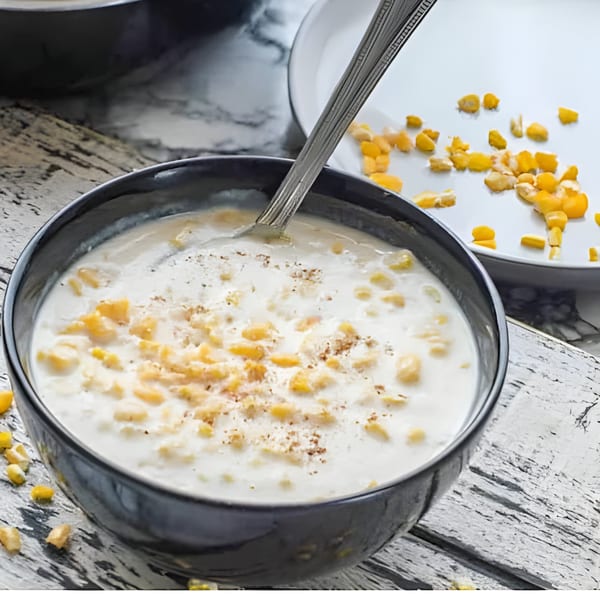 A black bowl of creamy corn soup with a spoon, on a rustic white wooden surface.